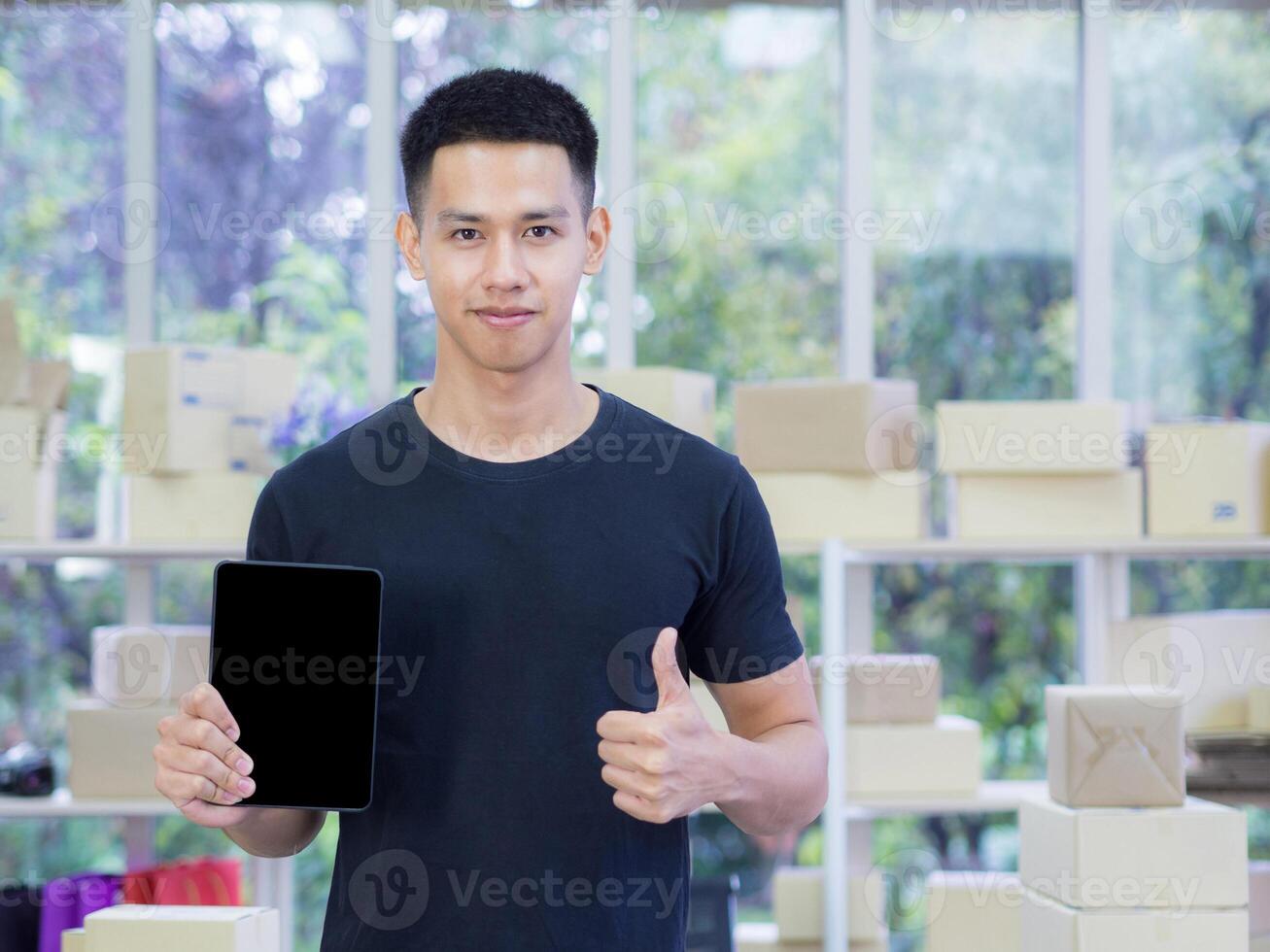 Young businessman wearing casual cloth holding a tablet, looking at the camera, and show fingers thumb up while standing in the office. Space for text. Concept of business and e-commerce photo