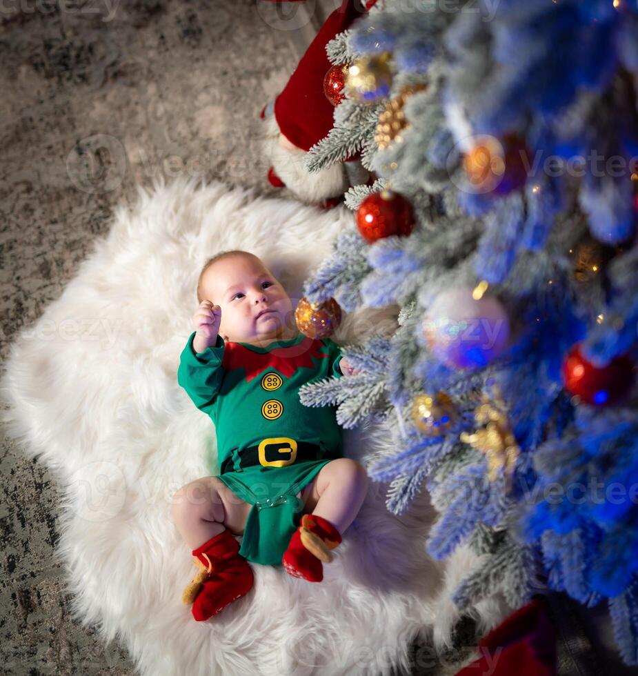 Top view of the sweet baby lying on the floor in a costume of elf. Baby near the fur tree at the new year. Side view. photo