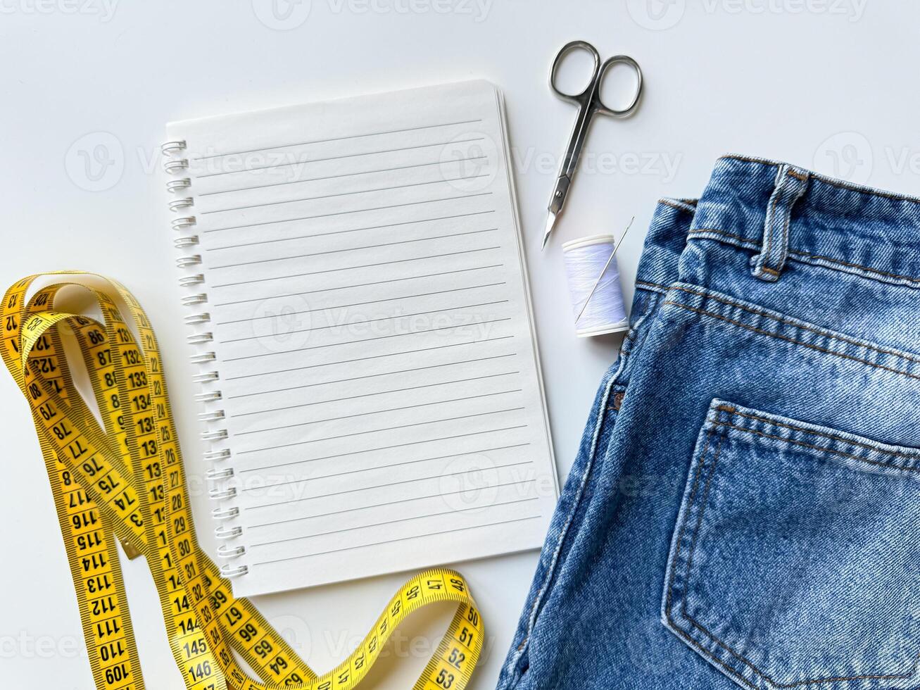 Tailoring and fashion design concept with denim jeans, yellow measuring tape, scissors, thread, and notepad with blank page for text on white background for planning and alterations. Top view. photo