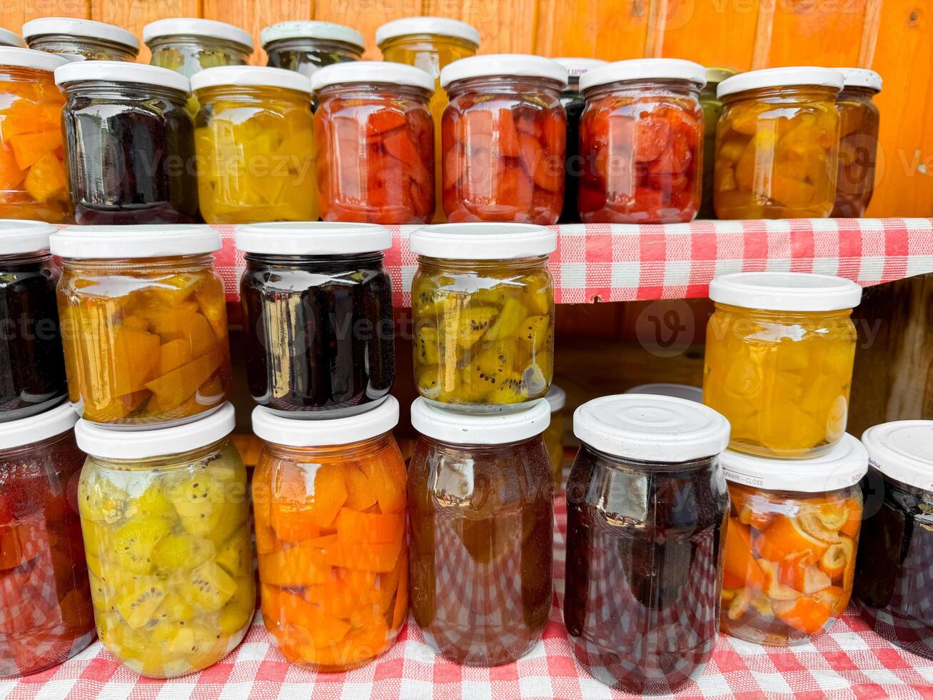 Assortment of homemade preserves in glass jars with checkered cloth, various pickled fruits and vegetables, colorful display, canned goods collection, close up with selective focus. photo