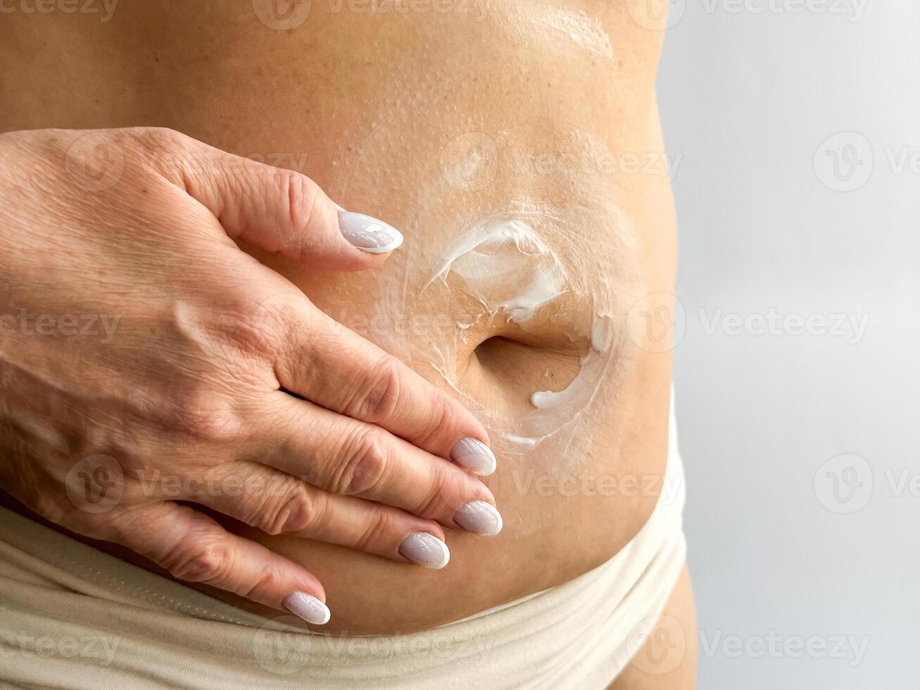 Close up of womans hand with white manicured nails applying cream to her stomach. Highlights skincare, body care, and personal hygiene, focusing on moisturizing and self care routines. photo