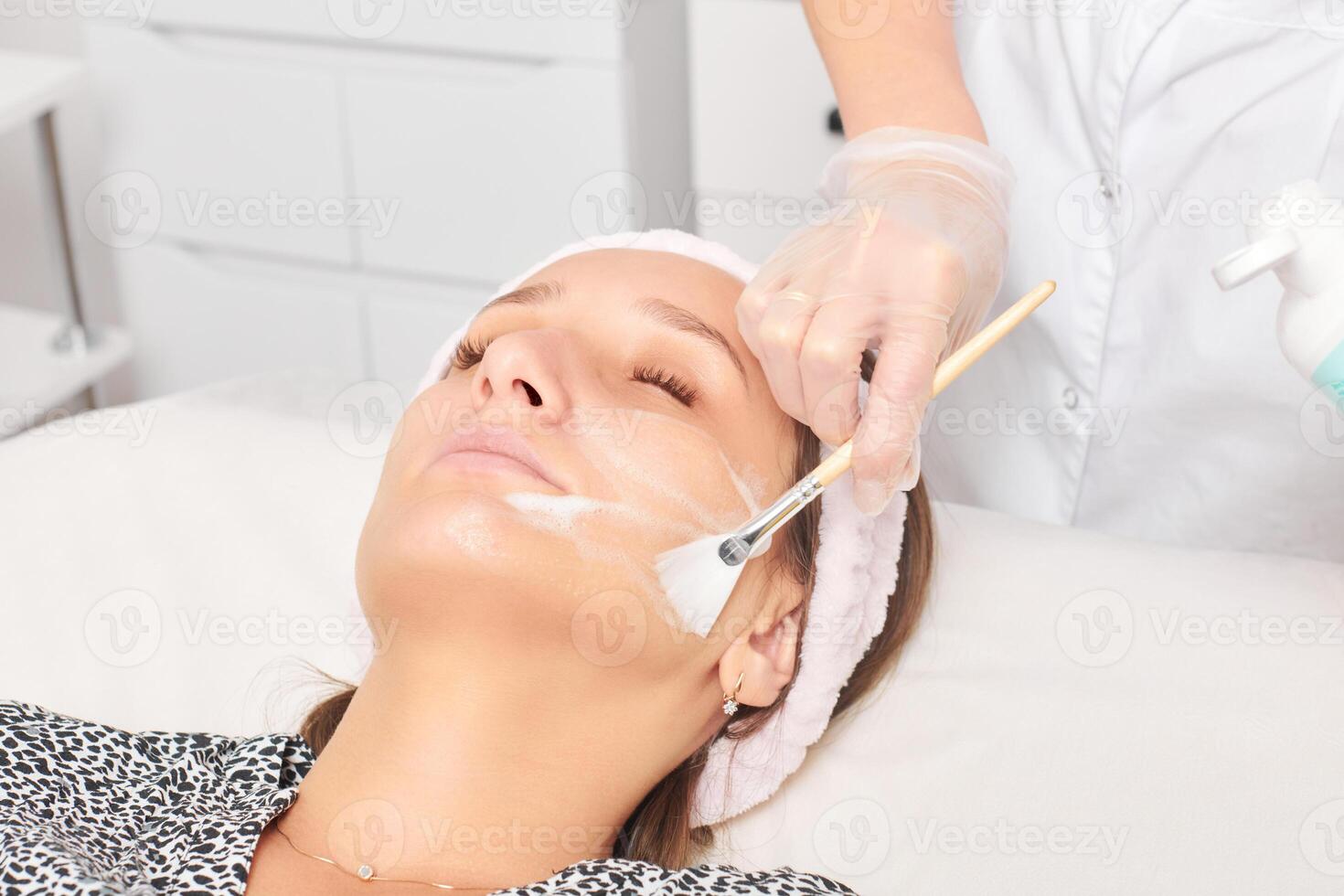 Beautician applying cosmetic cream mask on woman face for rejuvenation, procedure in beauty salon photo