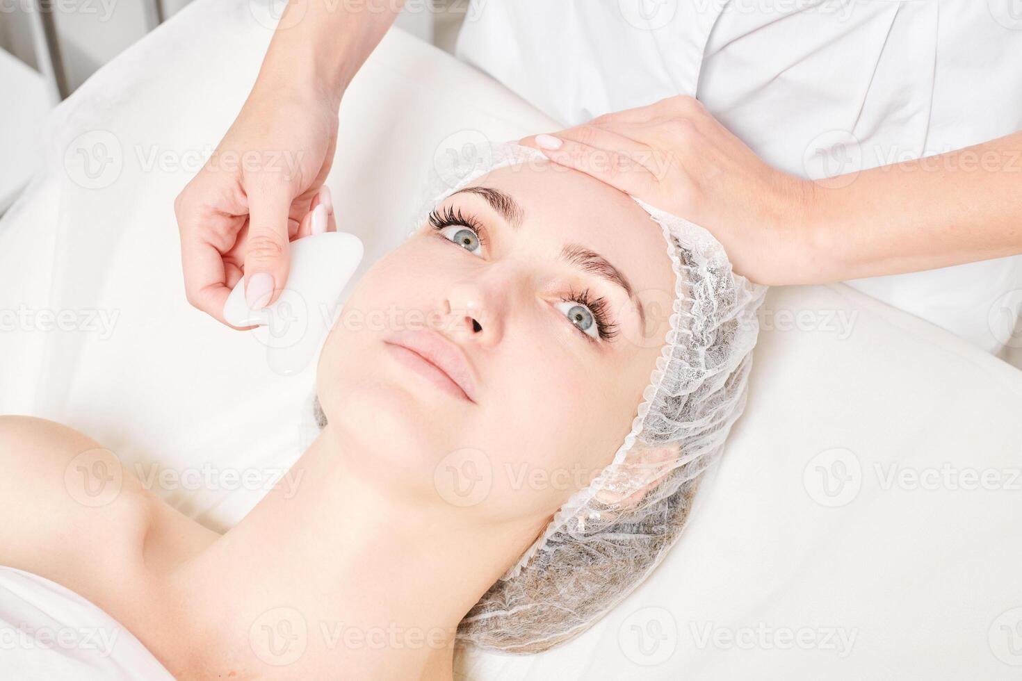 Cosmetologist making facial massage with Gua Sha stone of woman face skin for lymphatic drainage photo