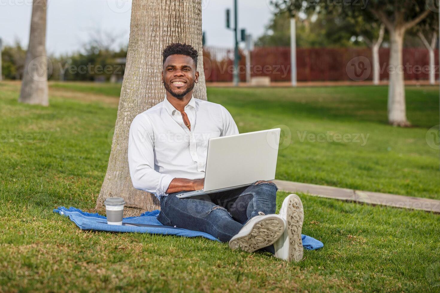 A smiling African-American man sitting under a tree, working or studying with a laptop computer. photo