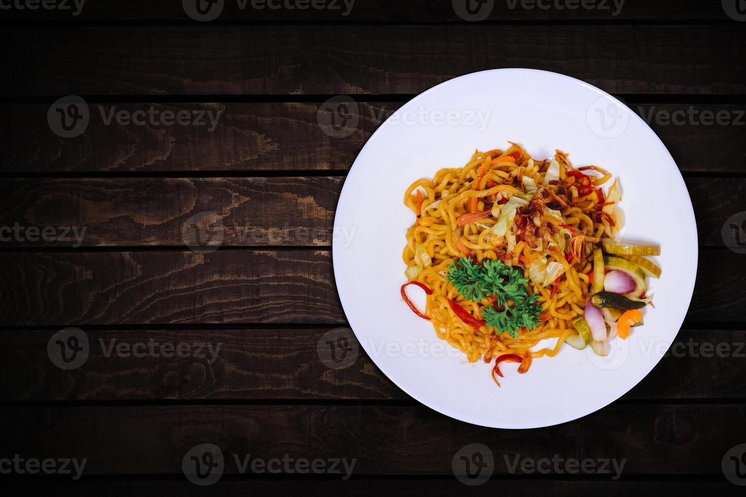 Spicy fried noodles with various additions on white plate on dark wooden table. top view. photo