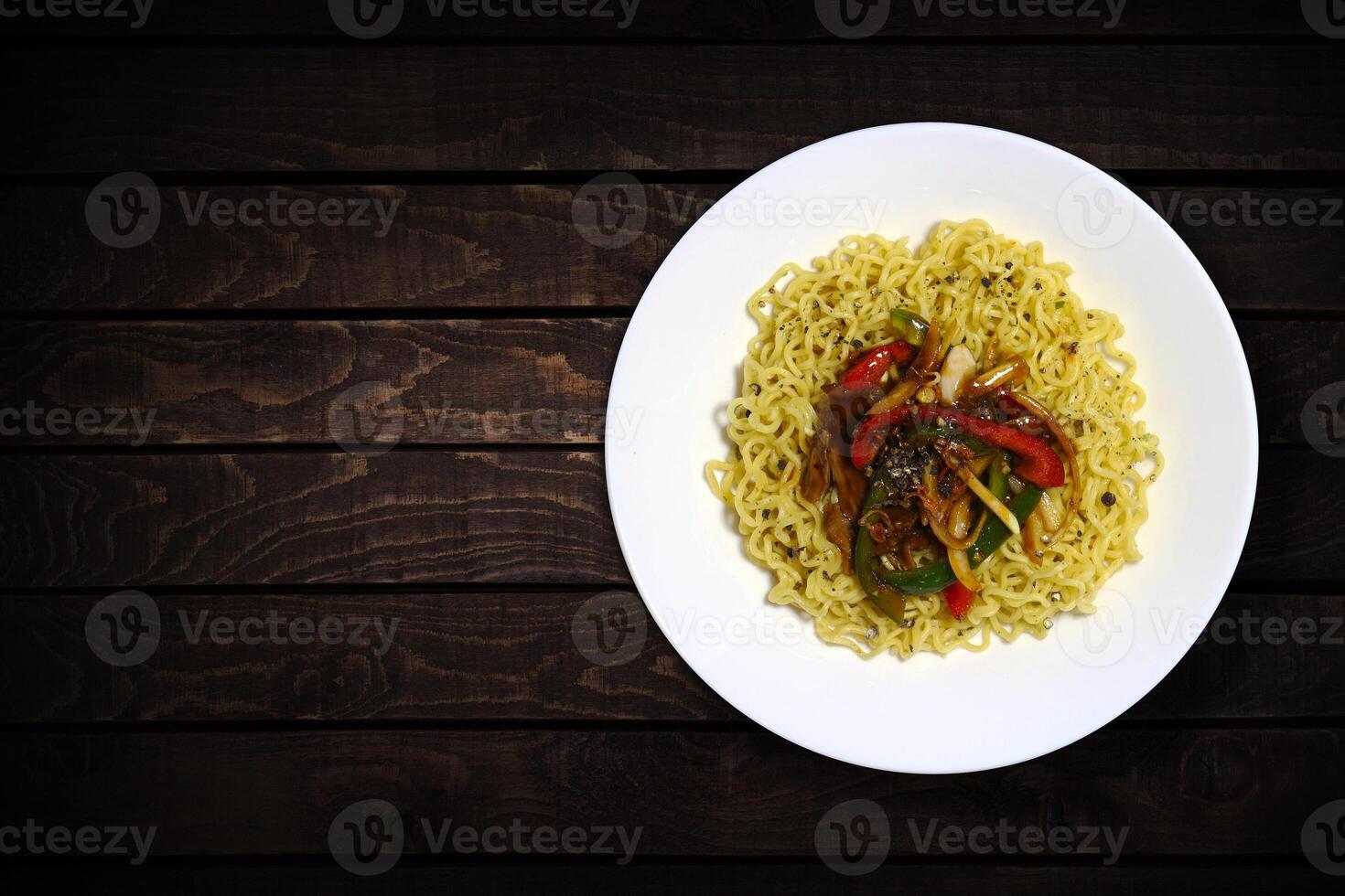Top view of instant noodles in white bowl with added chopped vegetables and black peppers on dark wooden background. photo