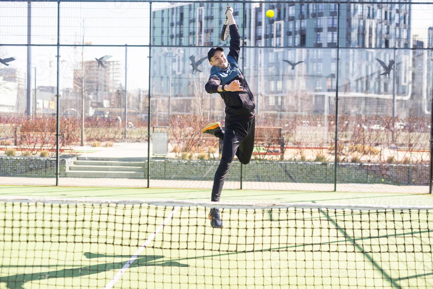 A padel player jump to the ball, good looking for posts and poster. Man with black racket playing a match in the open behind the net court outdoors. Professional sport concept with space for text photo