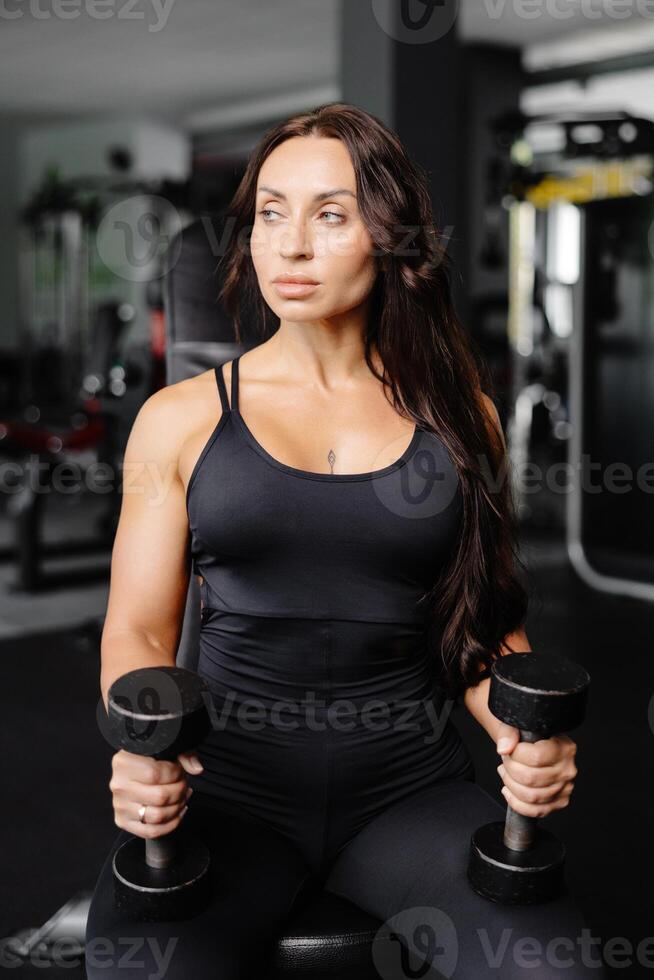 Young fit woman practicing dumbbell press while sitting on a bench. Slim female in sportswear exercises with weights in a gym photo