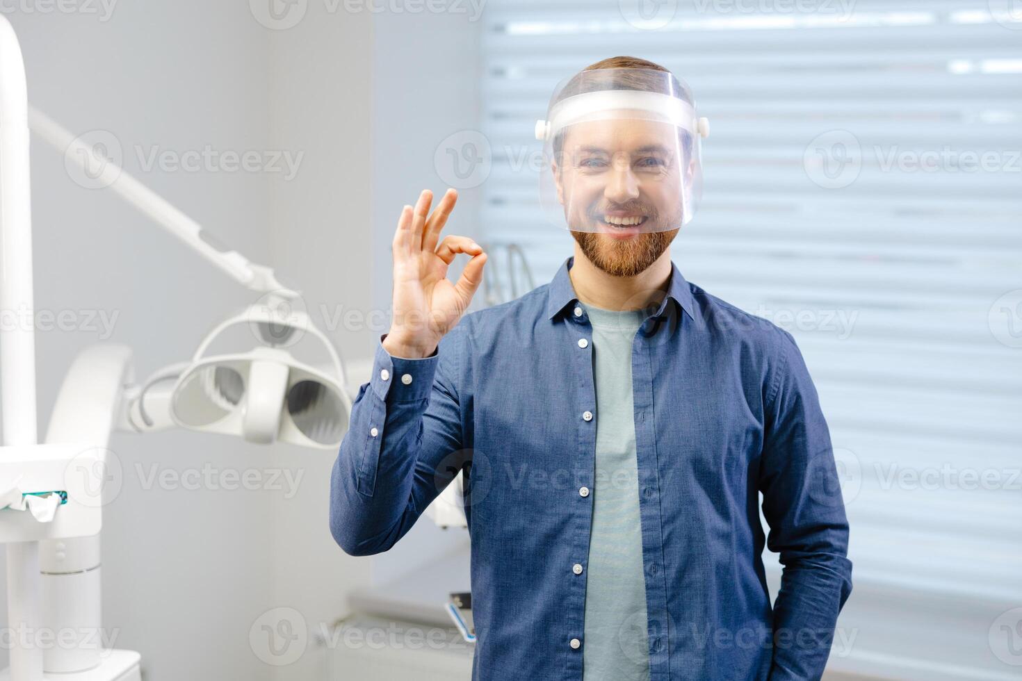 Portrait of handsome male dentist wearing in big protection medical shield on face preparing for dental surgery. Man smiling and looking at camera. photo