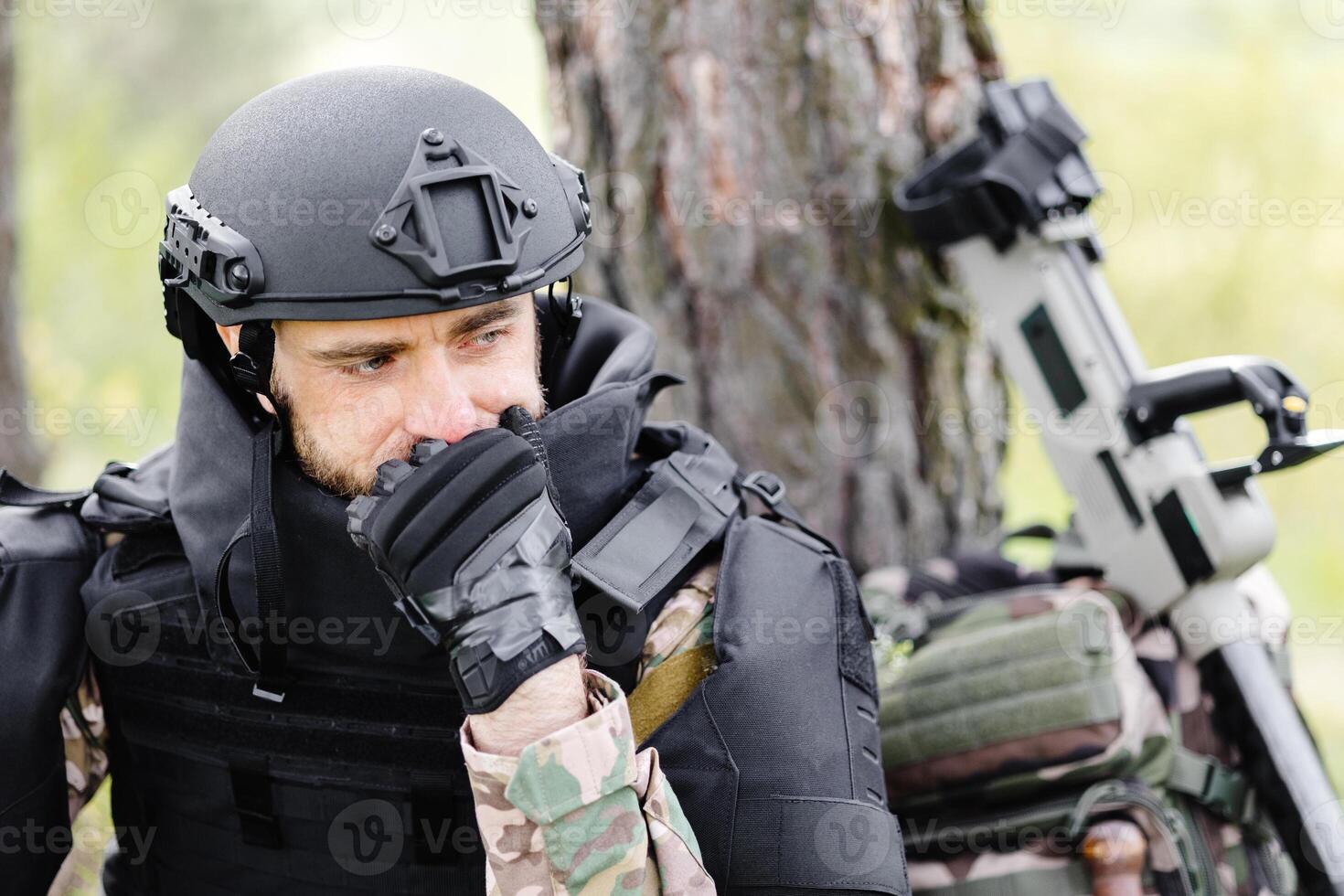 A man in a military uniform and a bulletproof vest sits in the forest near a metal detector and a military backpack. A man pauses in the work of demining the territory photo