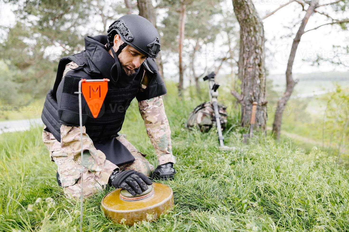A man in a military uniform and a bulletproof vest works in the forest to demine the territory. A man warns of danger by making a red mark photo