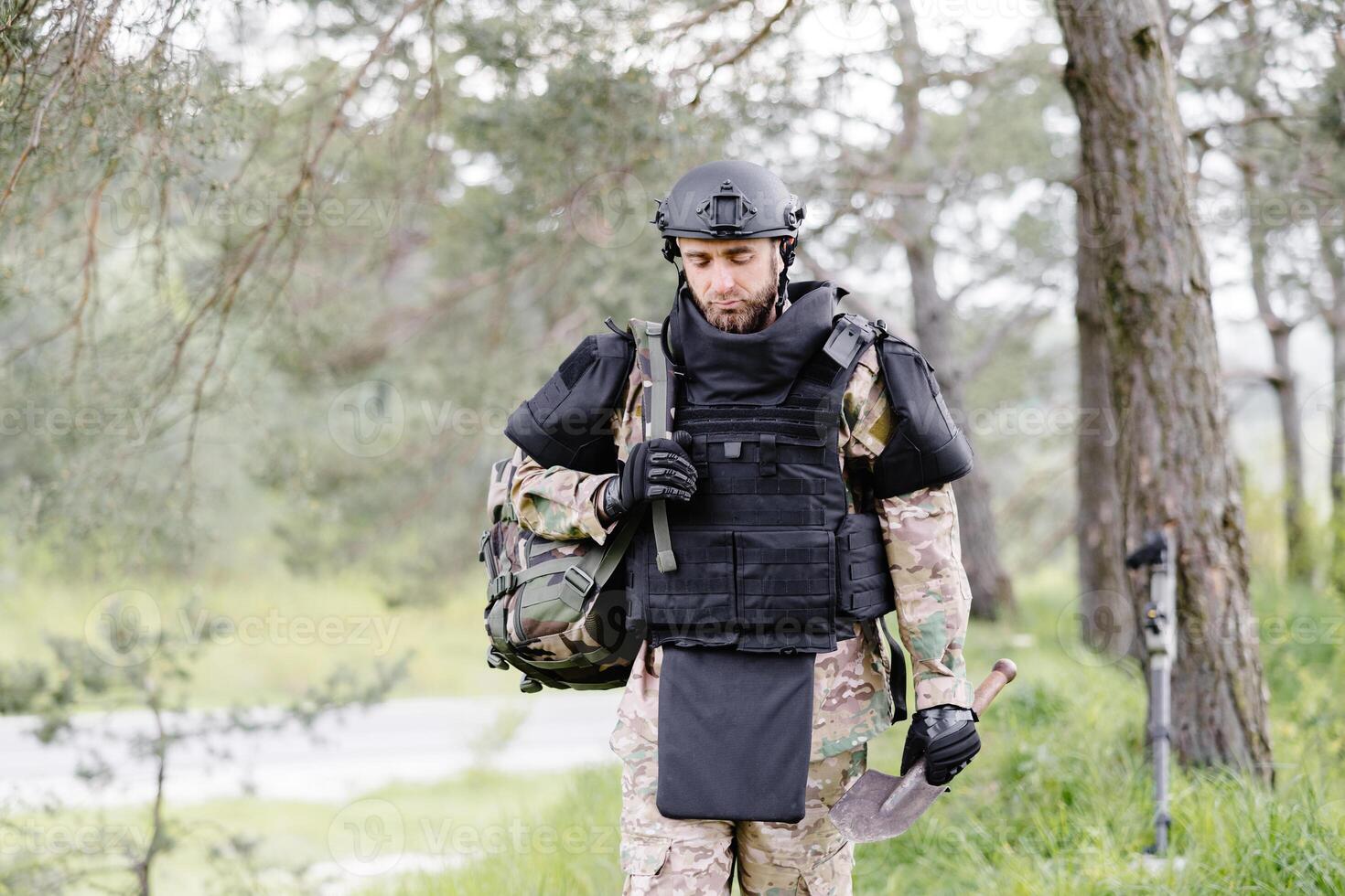 Young soldier in uniforms and tactical vest works in the forest and prepares for action at a temporary forest base. A man does in the work of demining the territory photo