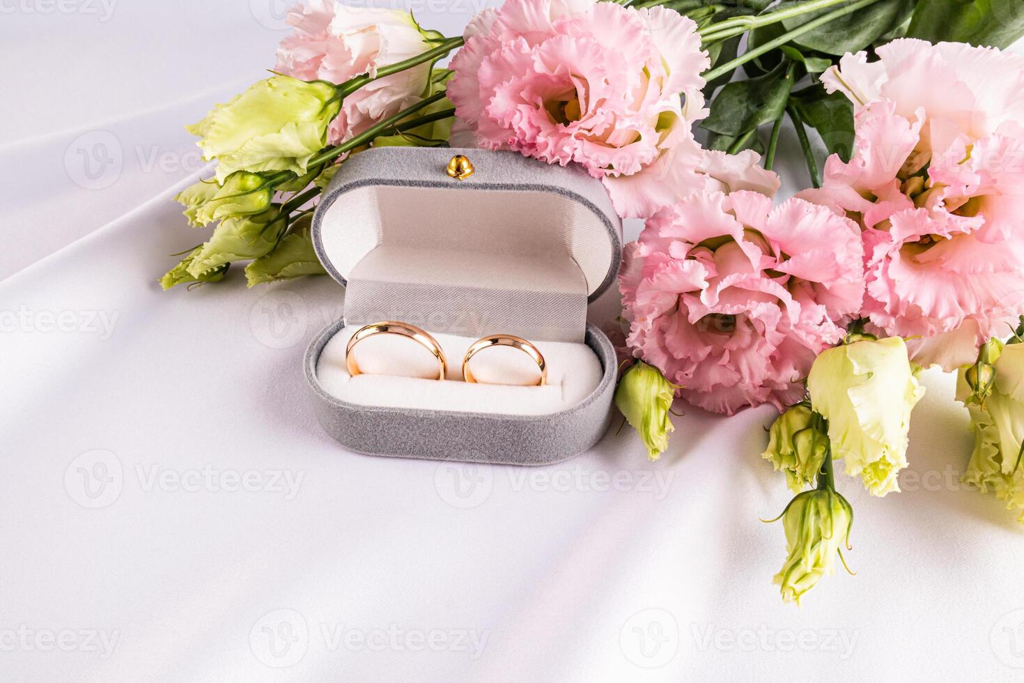 Two gold wedding classic rings in a gray velvet box on a white satin fabric with fresh pink flowers. Wedding concept. Front view photo