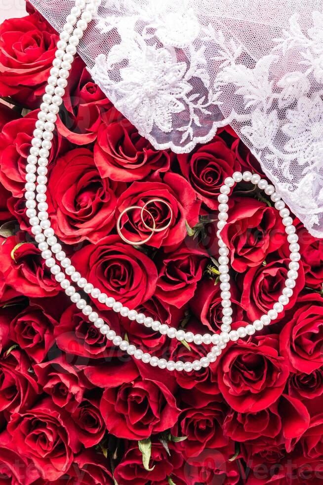 Wedding composition for the design of a postcard, cover, invitation. Pearl beads, two gold rings against a background of gorgeous red roses. Top view. photo