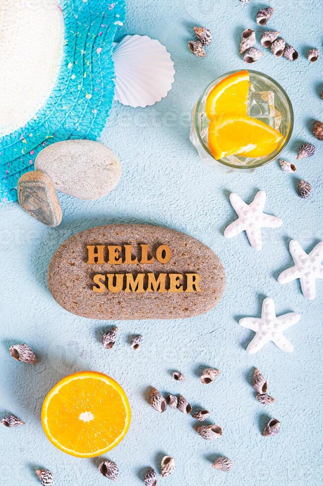 Hello summer text on stone, hat, cocktail, orange, starfish and seashells on blue top and vertical photo