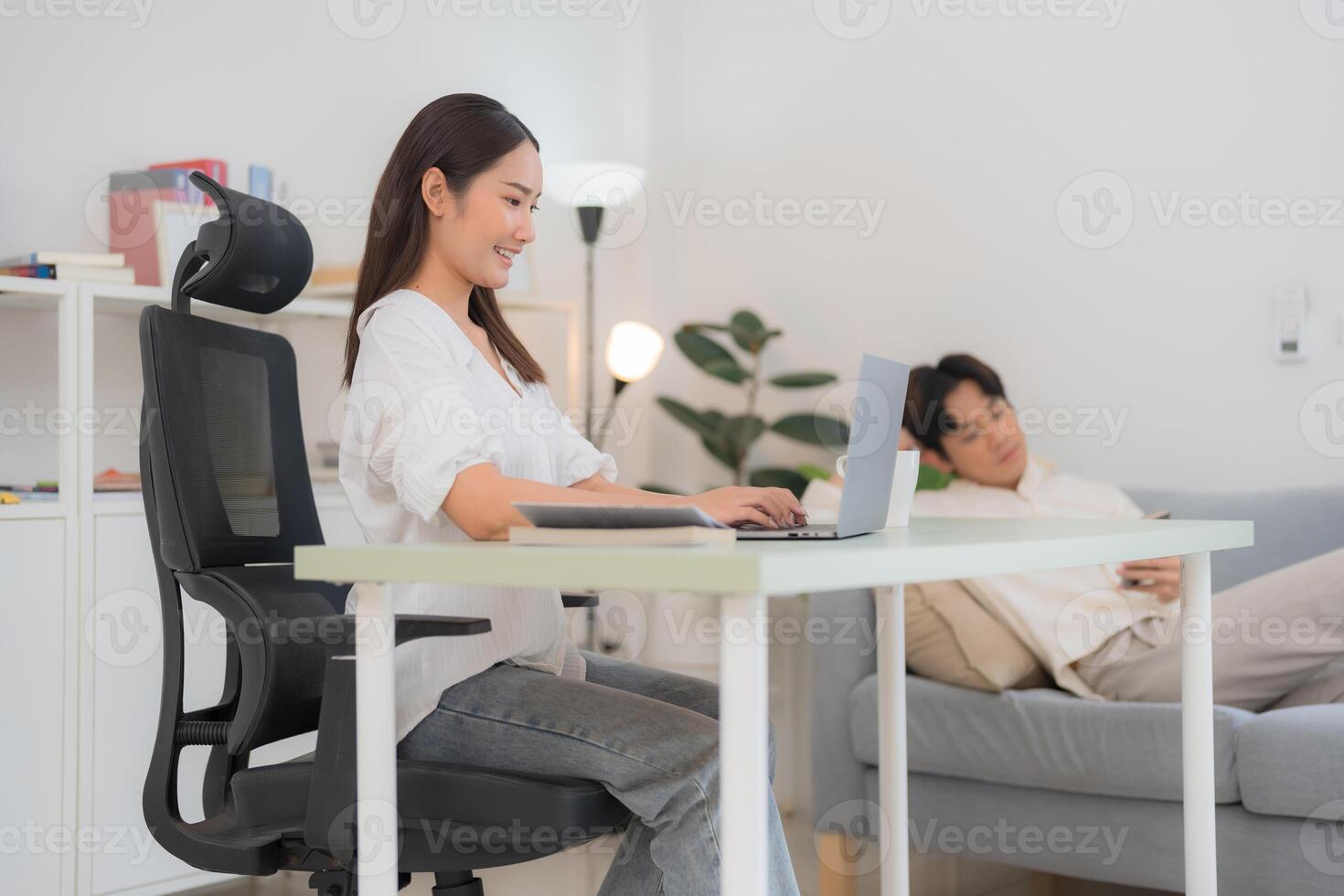 Young woman working on laptop with man resting on sofa in the background photo