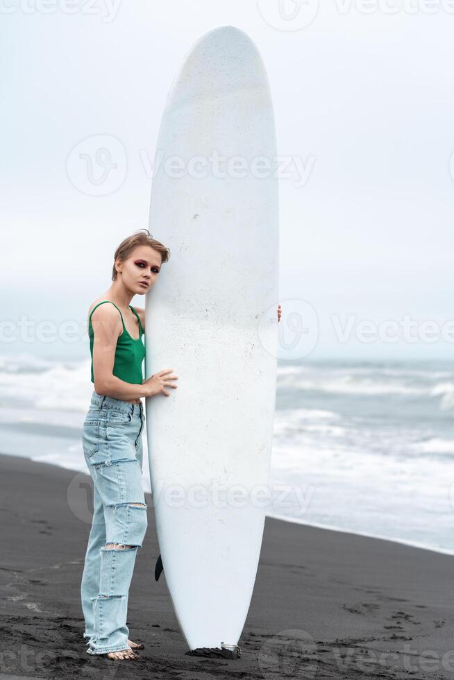 Woman surfer standing on beach, holding surfboard vertically on background of breaking ocean waves photo