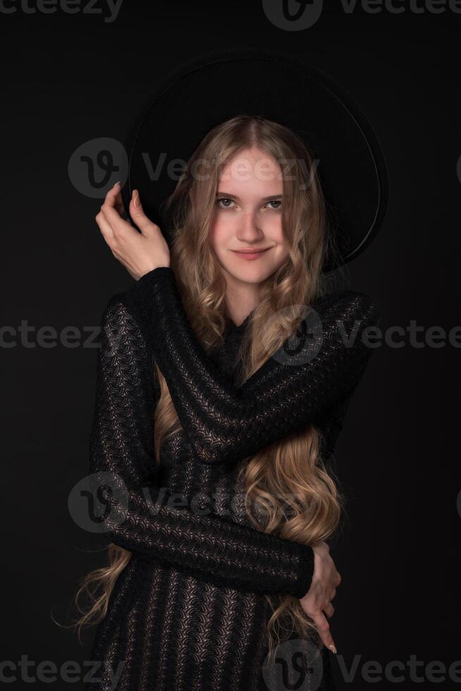 Woman with smirk on lips holds brim of hat with one hand, placing other on waist, looking at camera photo