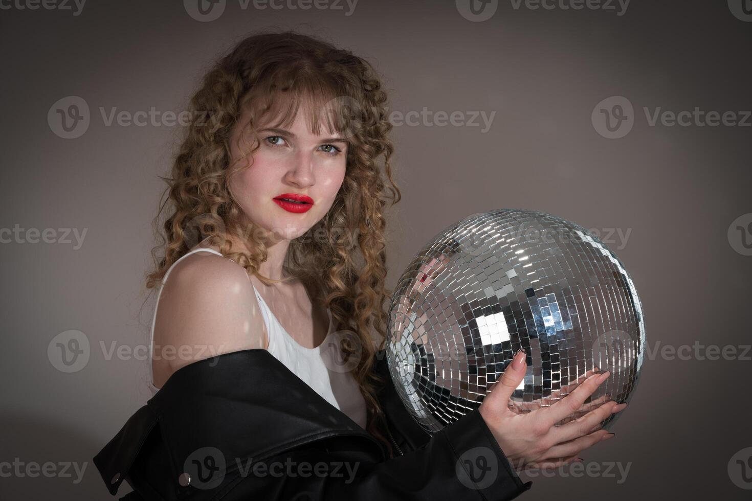 Woman holds mirror disco ball, looking at camera during party in nightclub. Young adult pin up girl photo