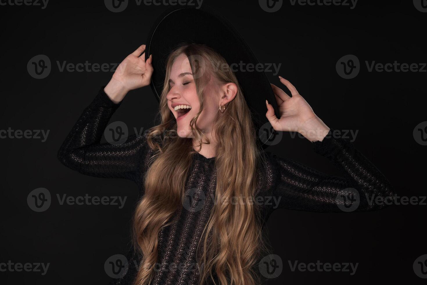 Woman with closed eyes and toothy smile, turning head to side and holding brim of felt hat photo