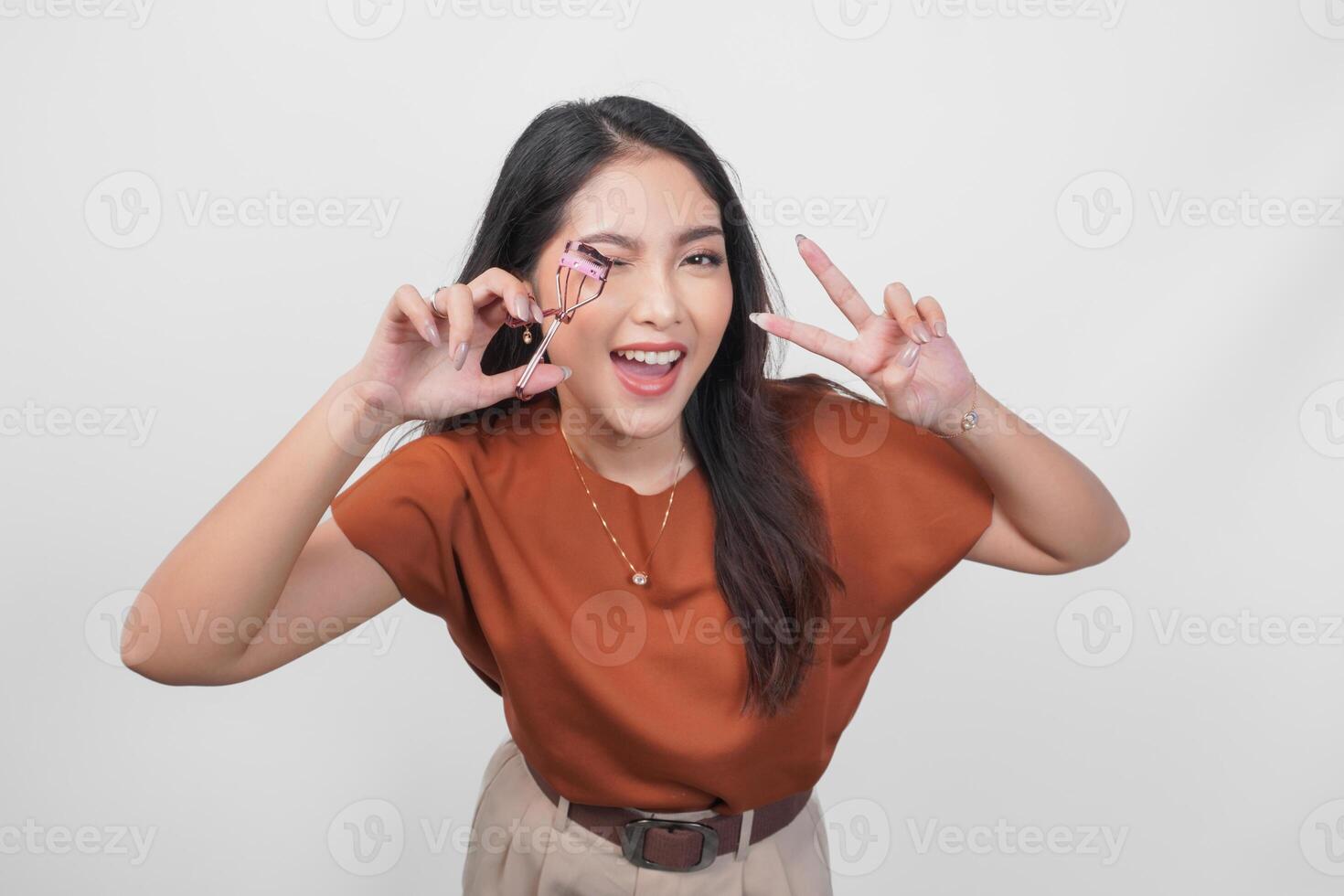 A happy young Asian woman is smiling and holding eyelash curler while wearing brown shirt over white isolated background. photo