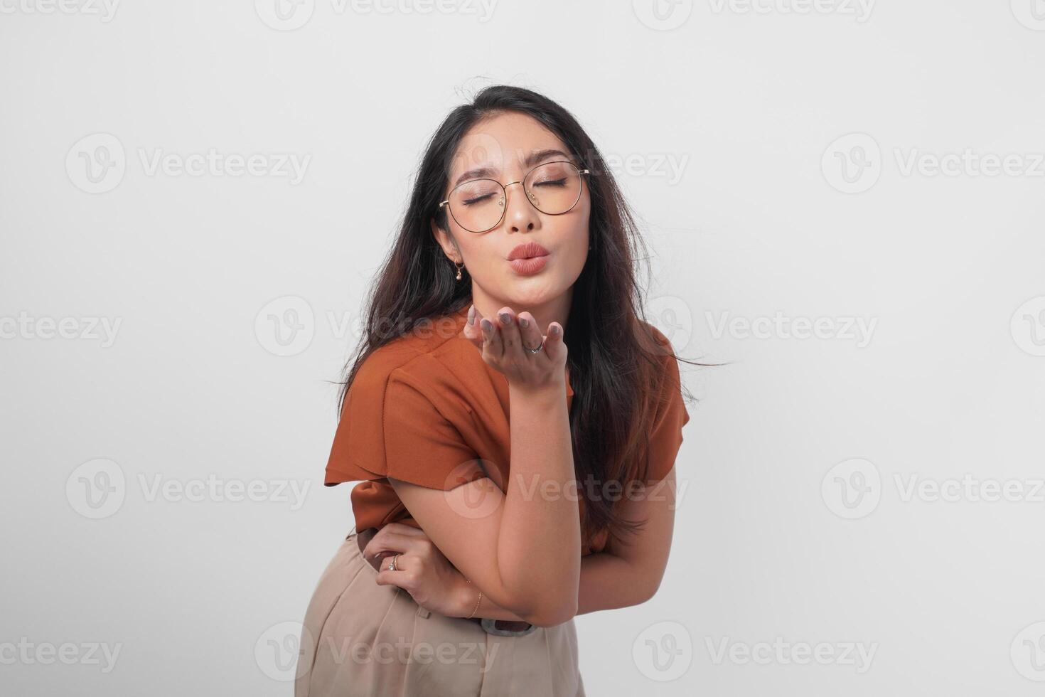 Cute Asian woman in brown shirt and eyeglasses sending blowing kiss with pout lips isolated on white background. photo
