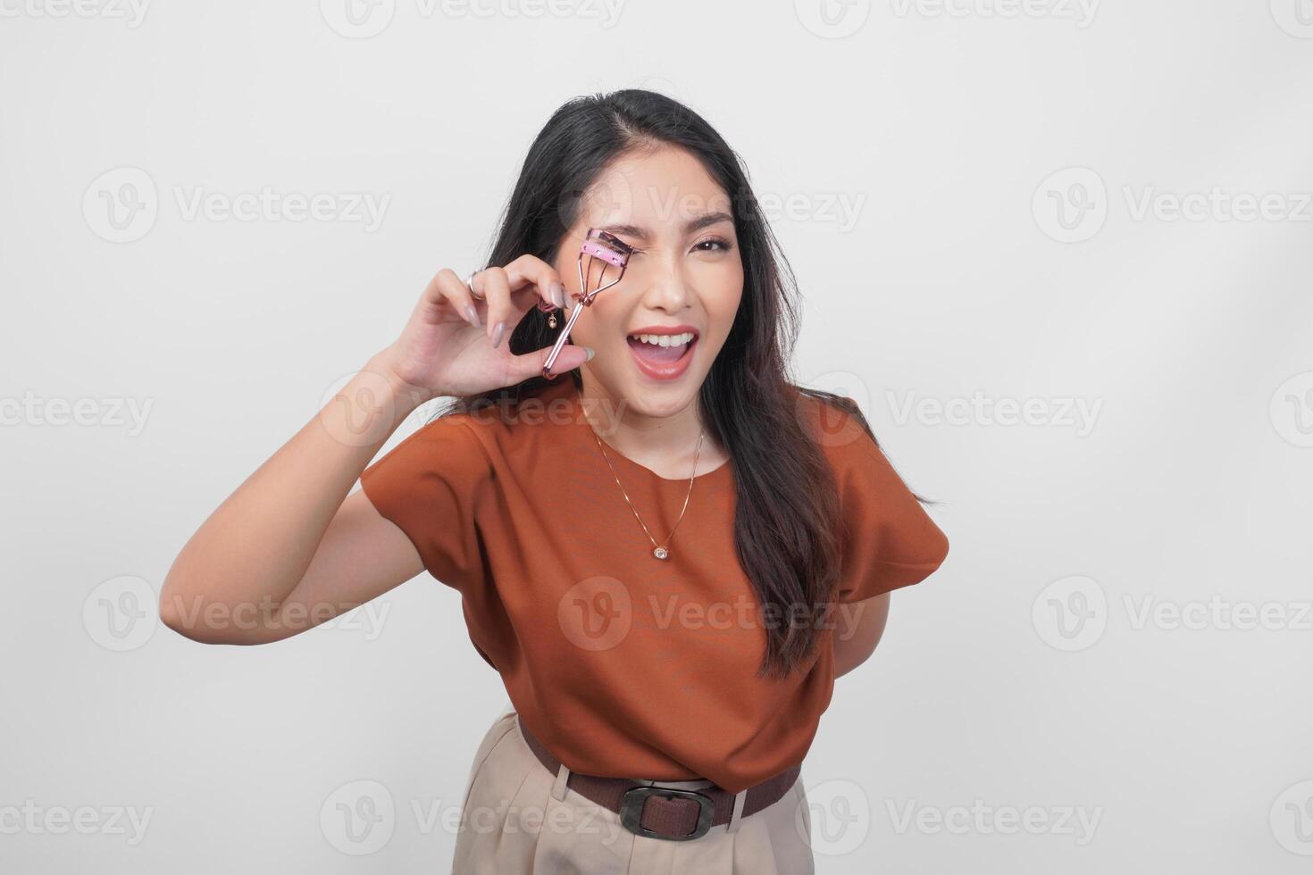 A happy young Asian woman is smiling and holding eyelash curler while wearing brown shirt over white isolated background. photo