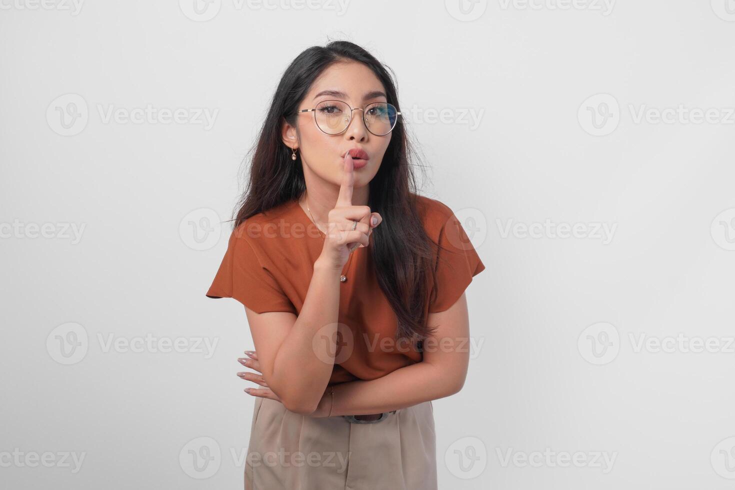 Young Asian woman wearing brown shirt and eyeglass looking to camera while putting a finger in front of lips gesturing stay silent or stay quiet, isolated by white background. photo