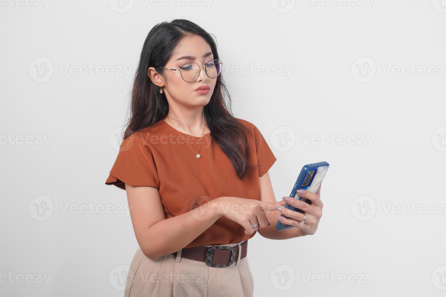 Thoughtful young Asian woman wearing brown shirt and eyeglasses looking aside while holding smartphone isolated over white background. photo