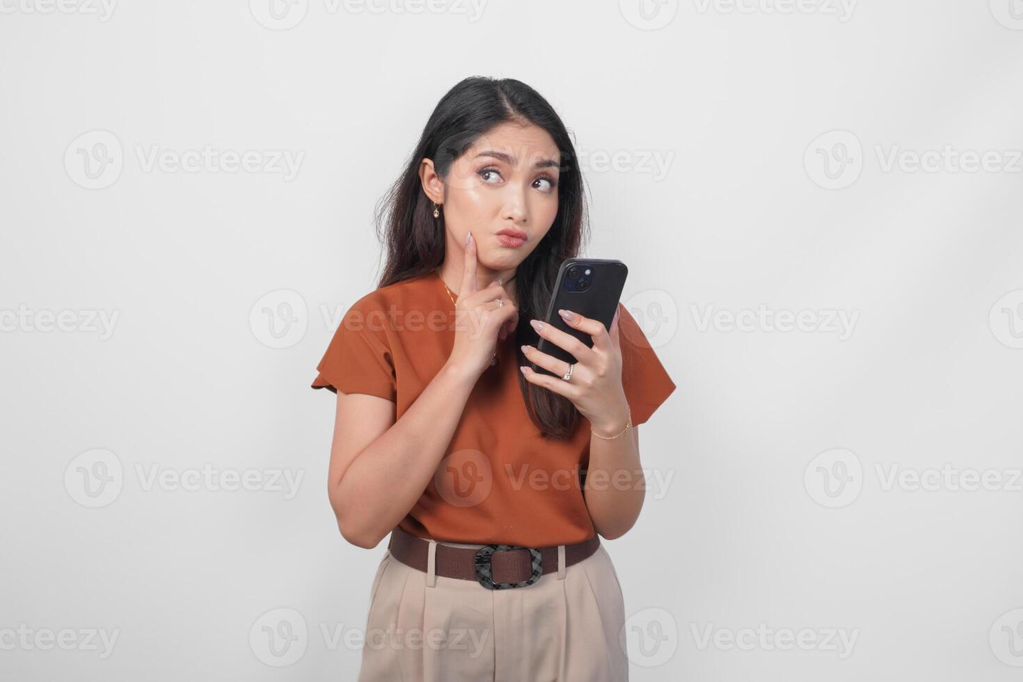 Thoughtful young woman wearing brown shirt looking aside while holding smartphone isolated over white background. photo