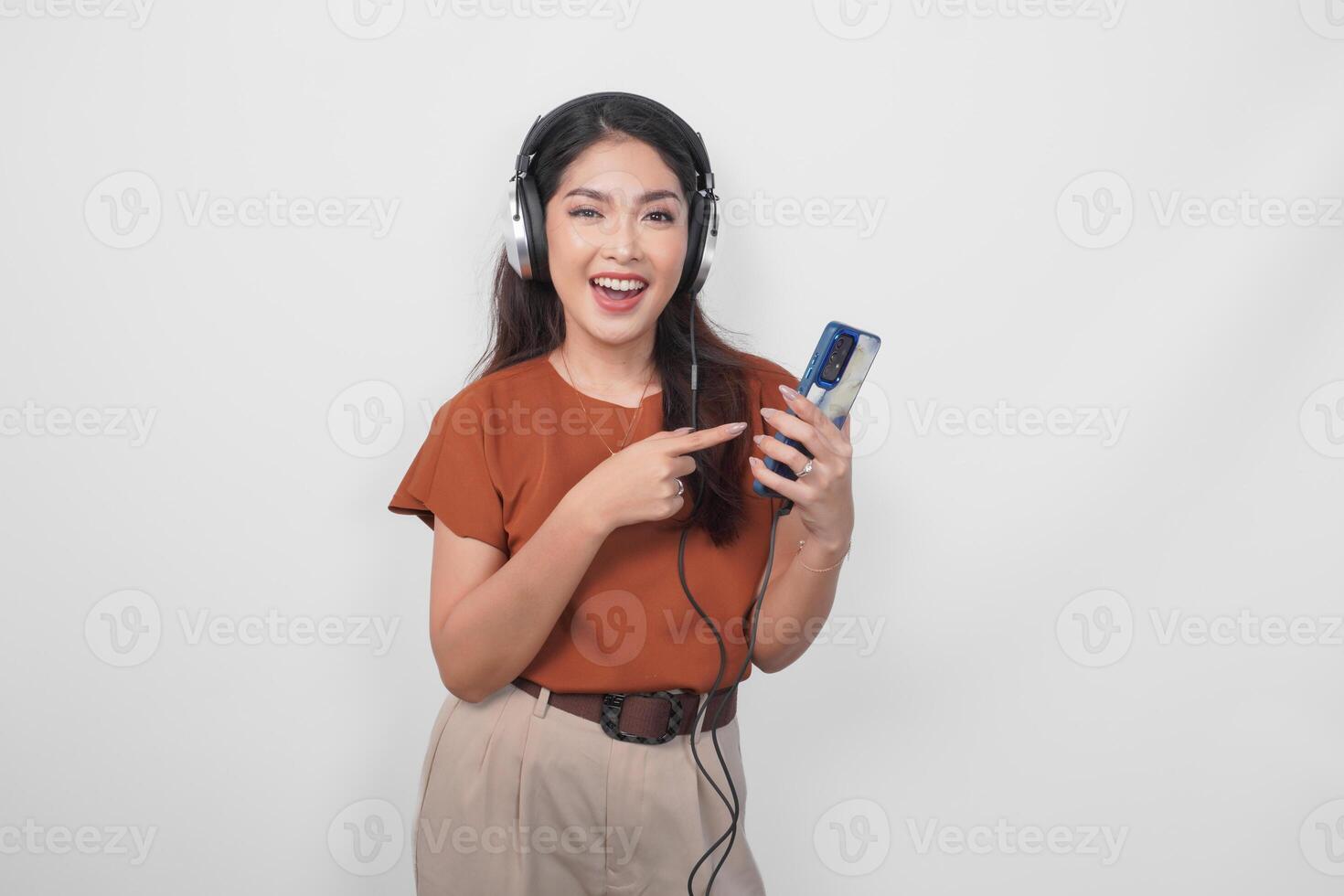 Attractive young Asian woman in brown shirt pointing to the smartphone and listening to music using wireless headphone. photo