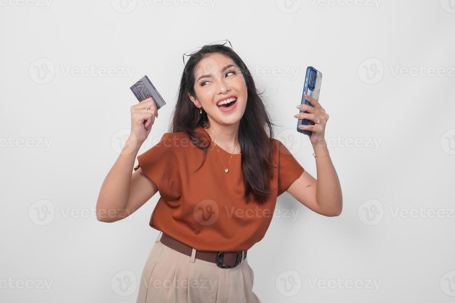 Happy successful Asian woman wearing brown shirt is holding her smartphone and credit card over isolated white background. photo