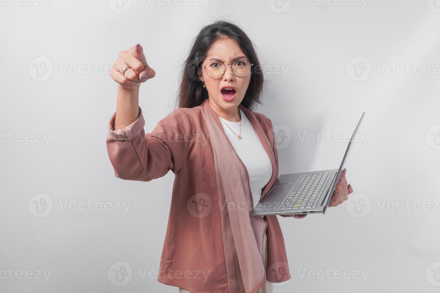 Enraged young Asian business woman holding a laptop pointing to the camera with angry expression, isolated by white background. photo