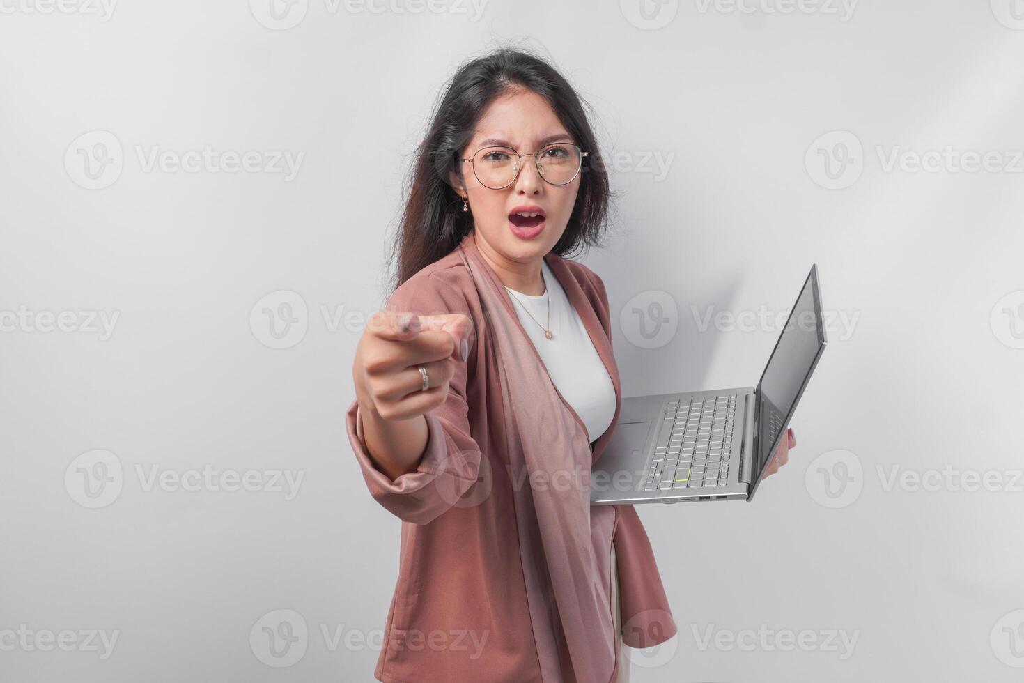 Enraged young Asian business woman holding a laptop pointing to the camera with angry expression, isolated by white background. photo