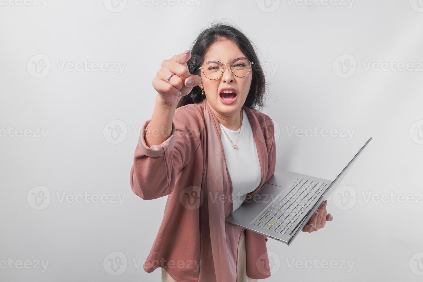Enraged young Asian business woman holding a laptop pointing to the camera with angry expression, isolated by white background. photo