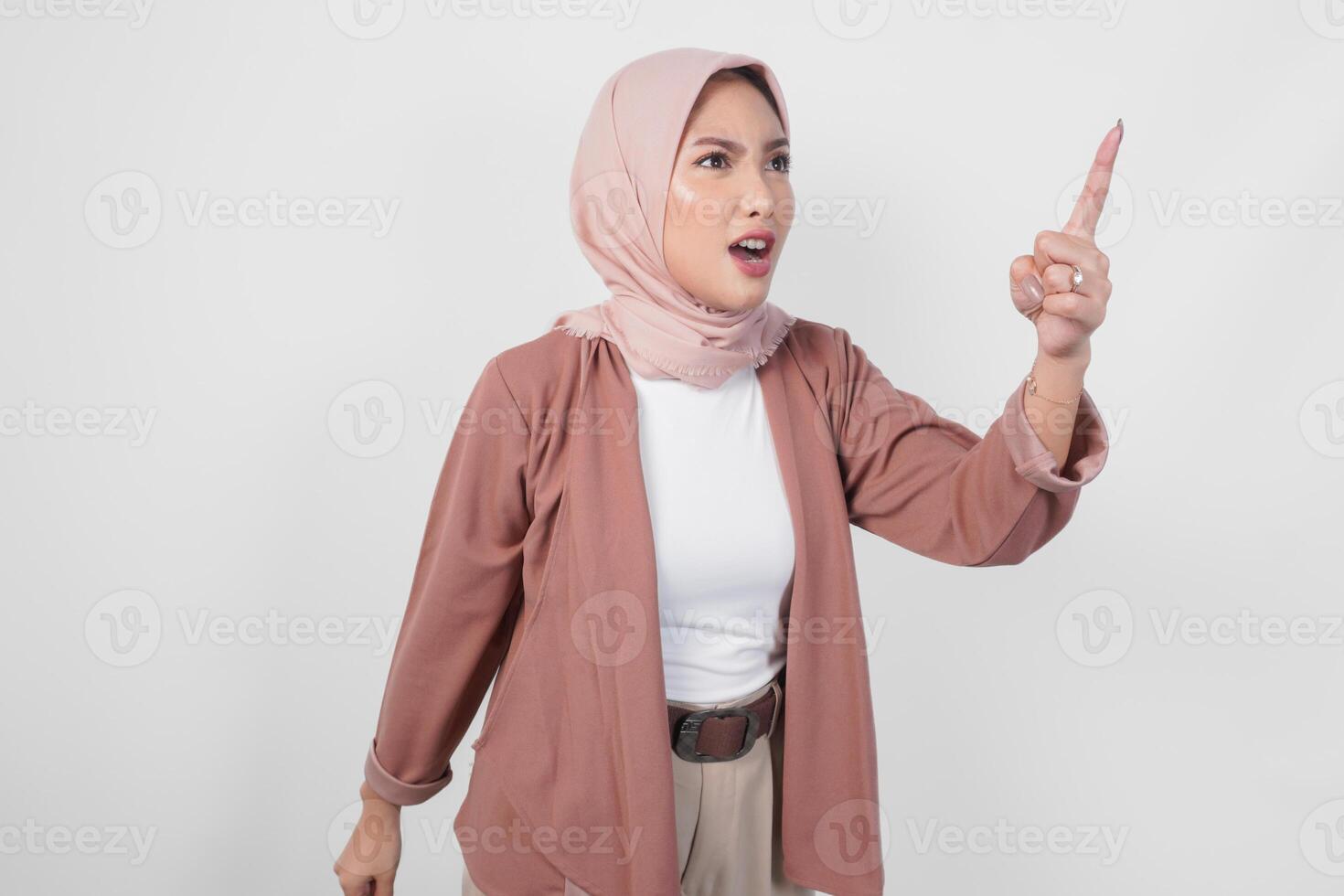 Young Asian Muslim woman wearing hijab looking disgruntled irritated expression while pointing to camera, isolated by white background. photo