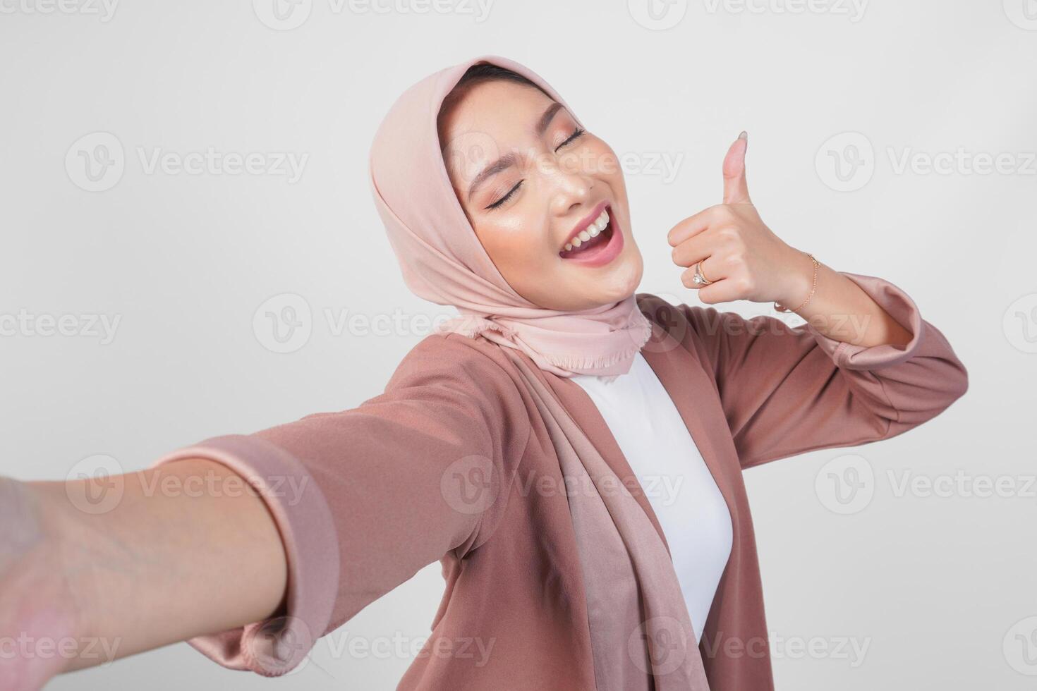 Smiling Asian woman in hijab posing for a selfie while giving thumbs up hand gesture of approval, isolated by white background. photo
