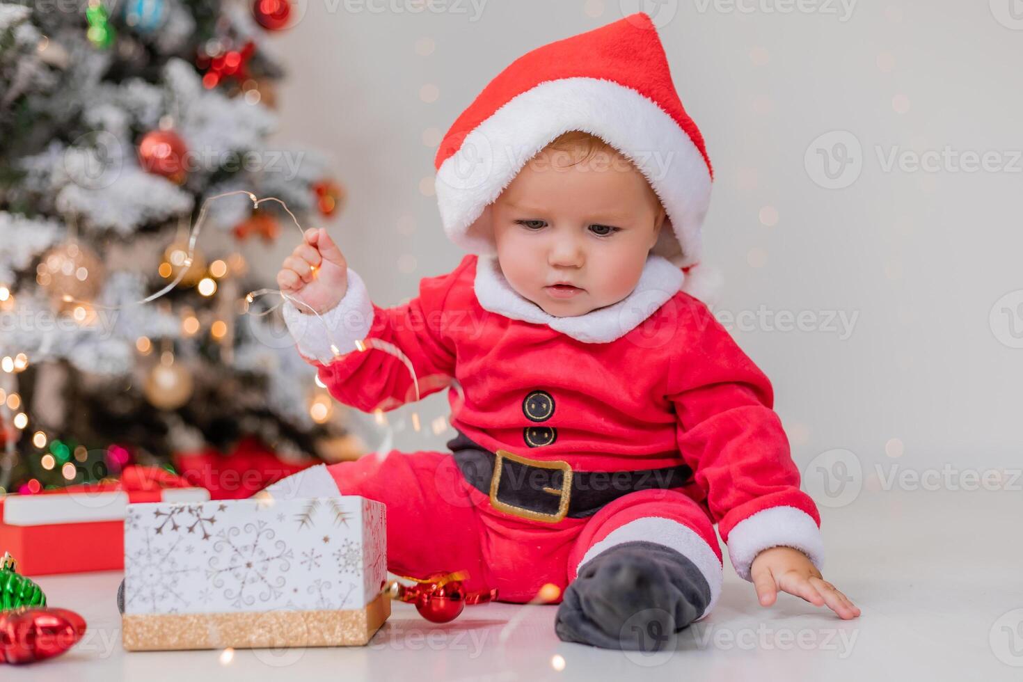 baby in a Santa costume is sitting next to the Christmas tree and playing with gift boxes. lifestyle photo