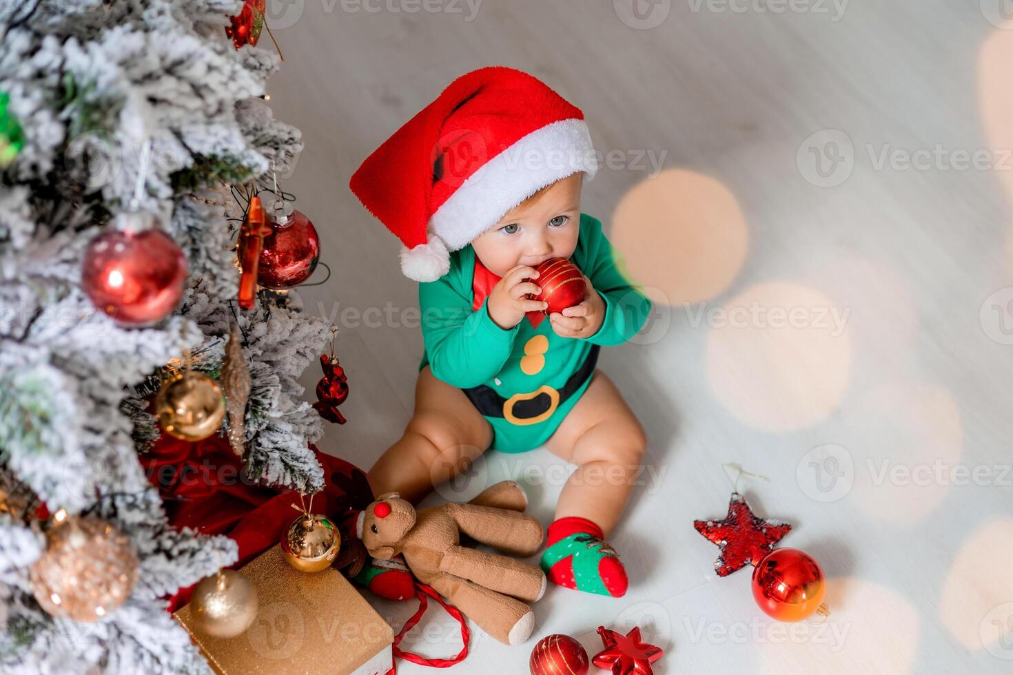 cute baby in a green bodysuit and a Santa hat decorates the Christmas tree with colorful balloons photo