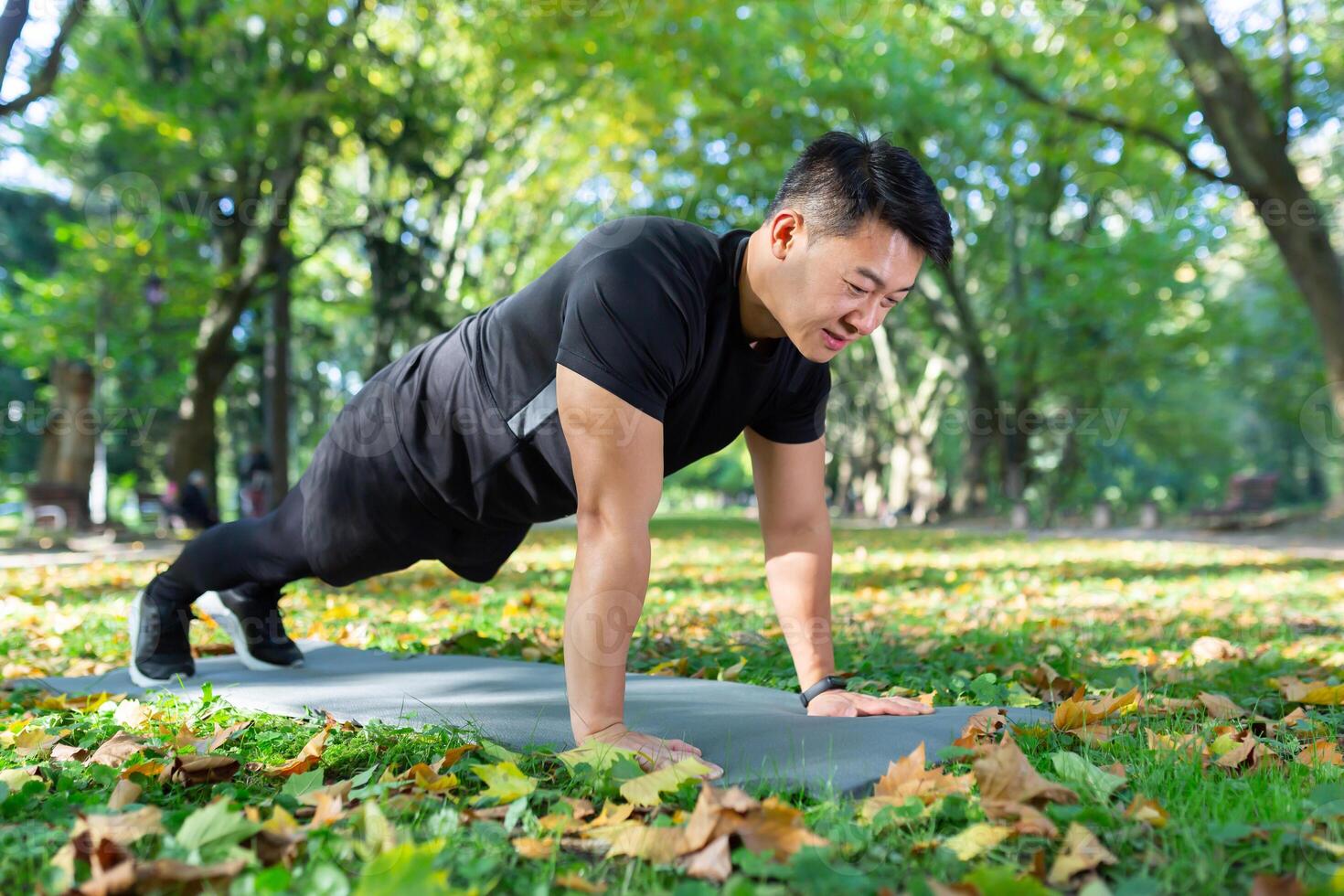 Asian athlete doing push-ups in park and doing plank on sports mat, man happy with workout on sunny day. photo