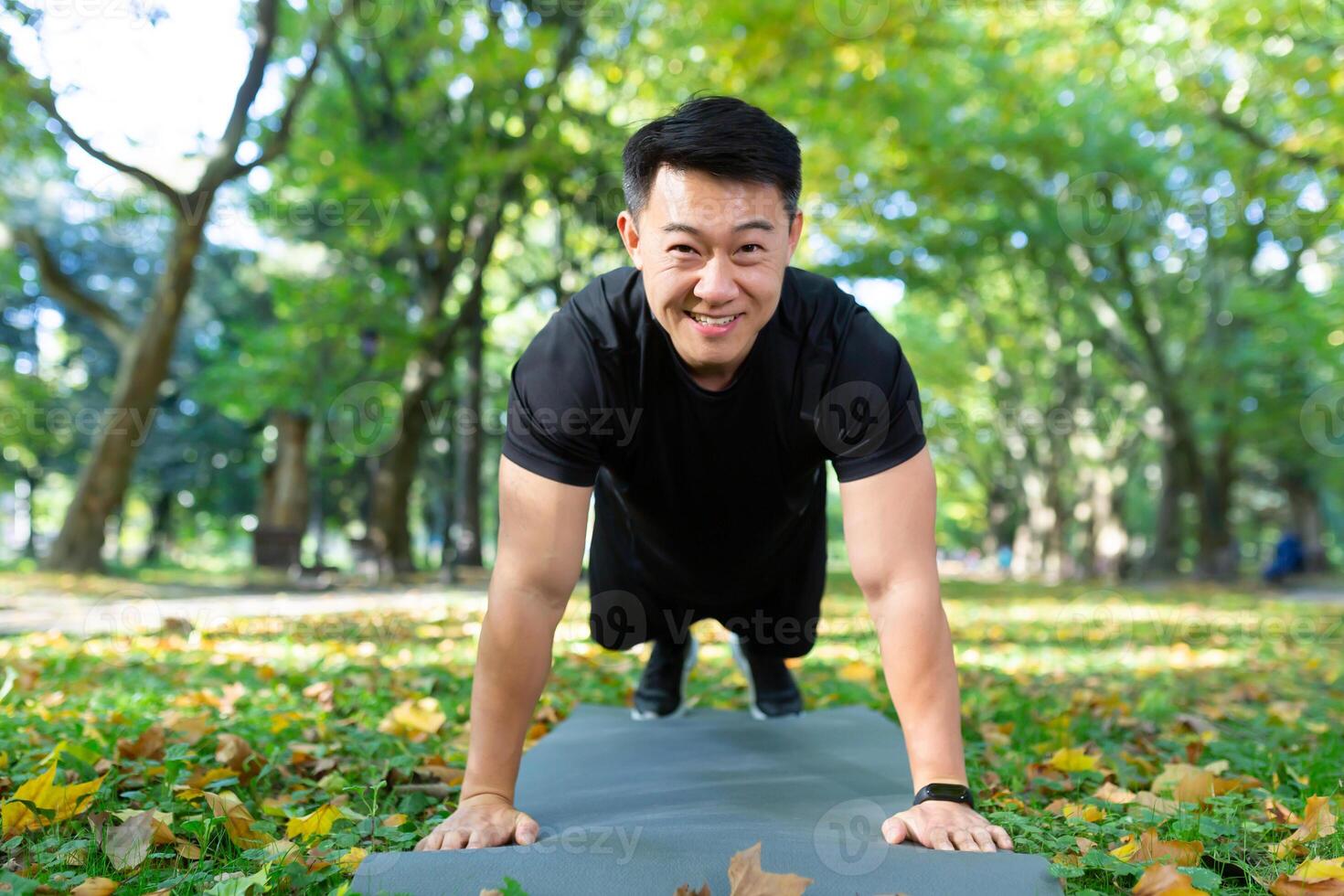 Portrait of happy male athlete, Asian looking at camera and smiling doing physical exercises, push-ups and plank in autumn park on sunny day. photo