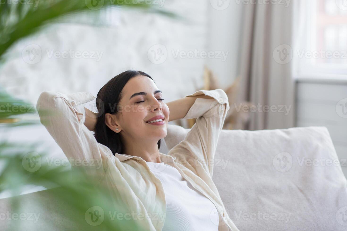 Close-up photo of a young beautiful woman relaxing at home with her eyes closed, her hands behind her head and daydreaming while sitting on the couch in the daytime