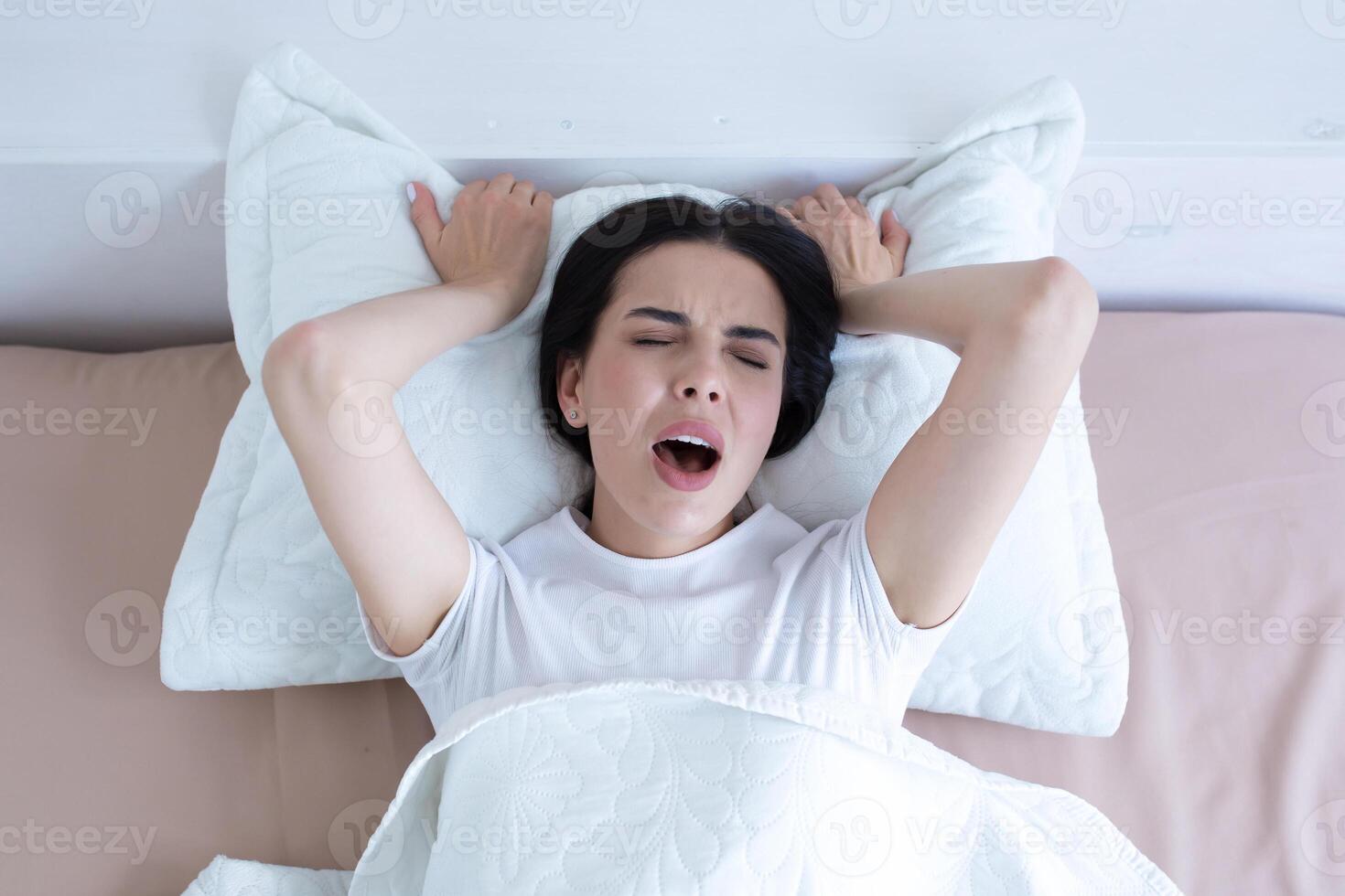 Frustrated and upset woman lying in bed alone at home crying and screaming, top view, brunette in white t-shirt pajamas photo