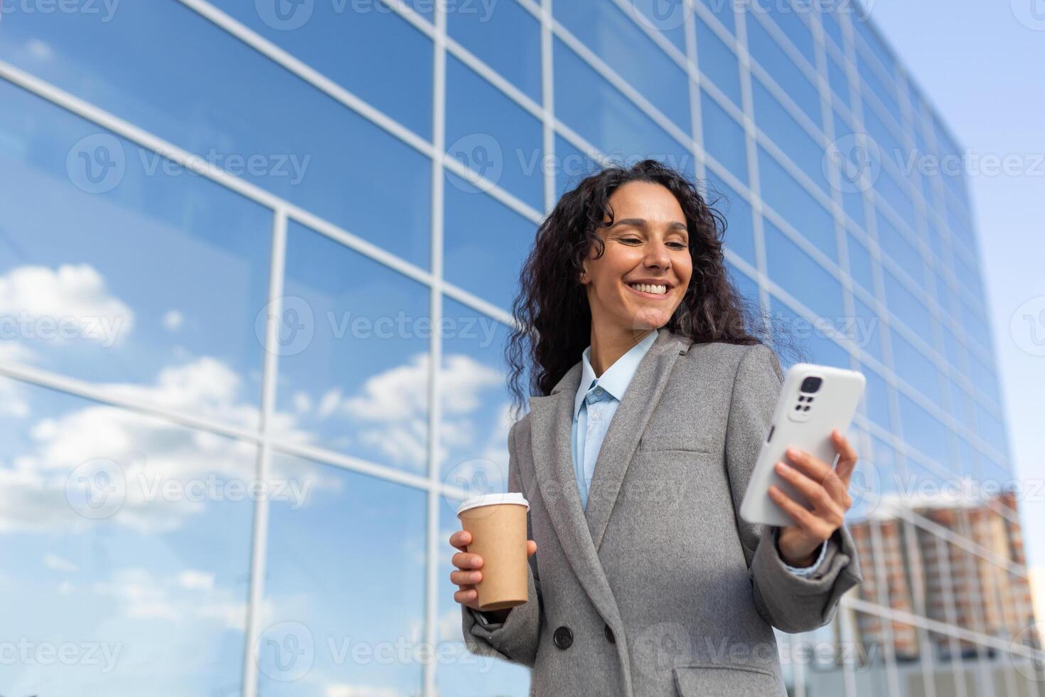 Successful business woman with smartphone goes to work in the morning in the office, hispanic woman with curly hair smiling and browsing online news outside with cup hot drink, in warm clothes coat. photo