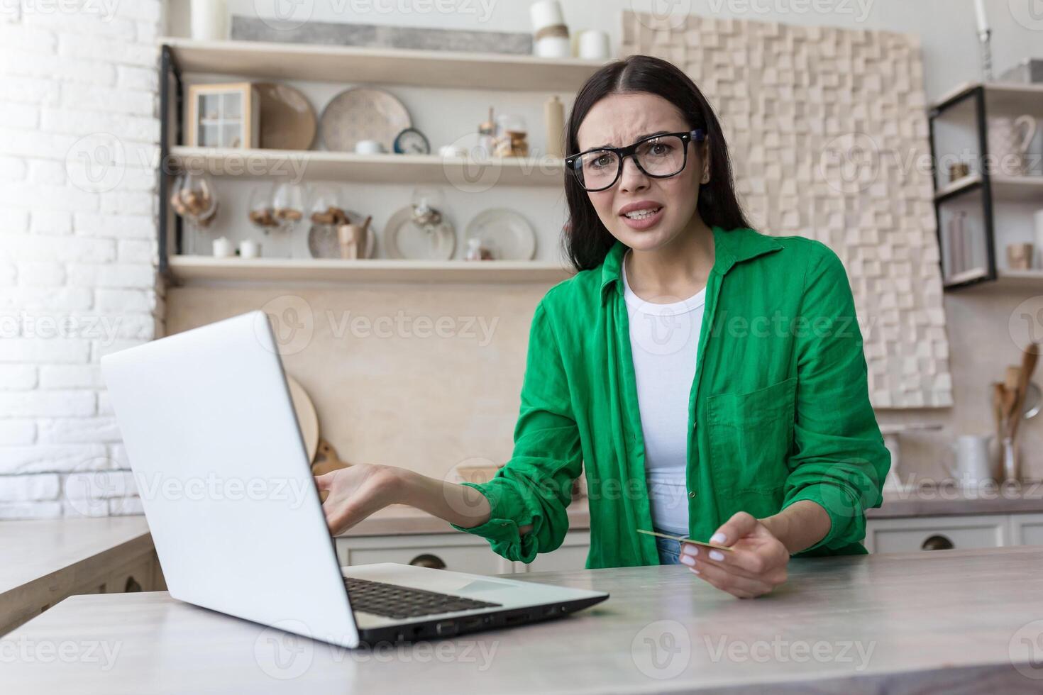 Angry young woman sitting at home in the kitchen, working on a laptop, having problems with a credit card, bill, payment. He spreads his hands and looks at the camera in confusion. photo