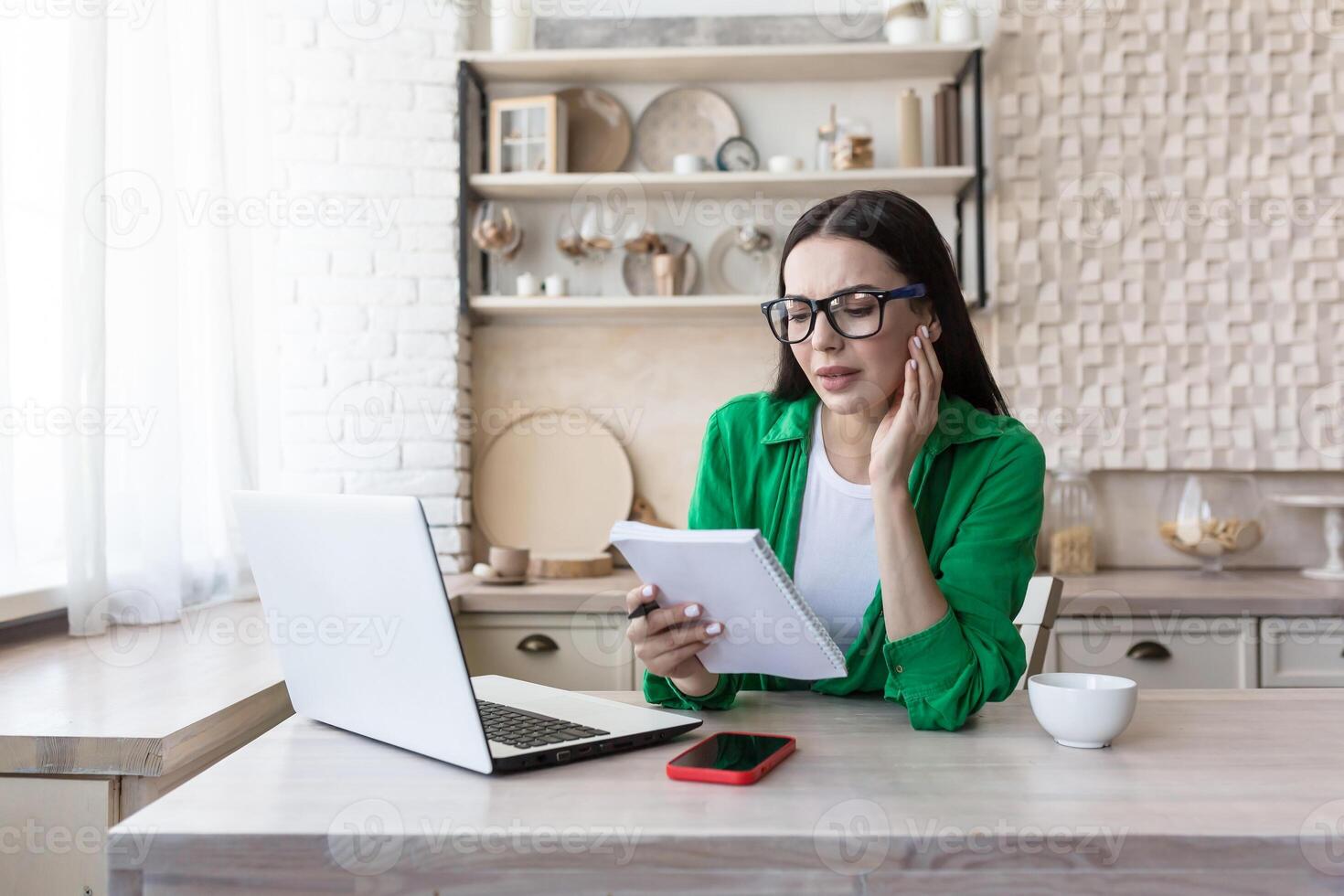 A young woman sits at home in the kitchen with a laptop, jots down the family budget in a notebook, counts bills, keeps a list of purchases. photo