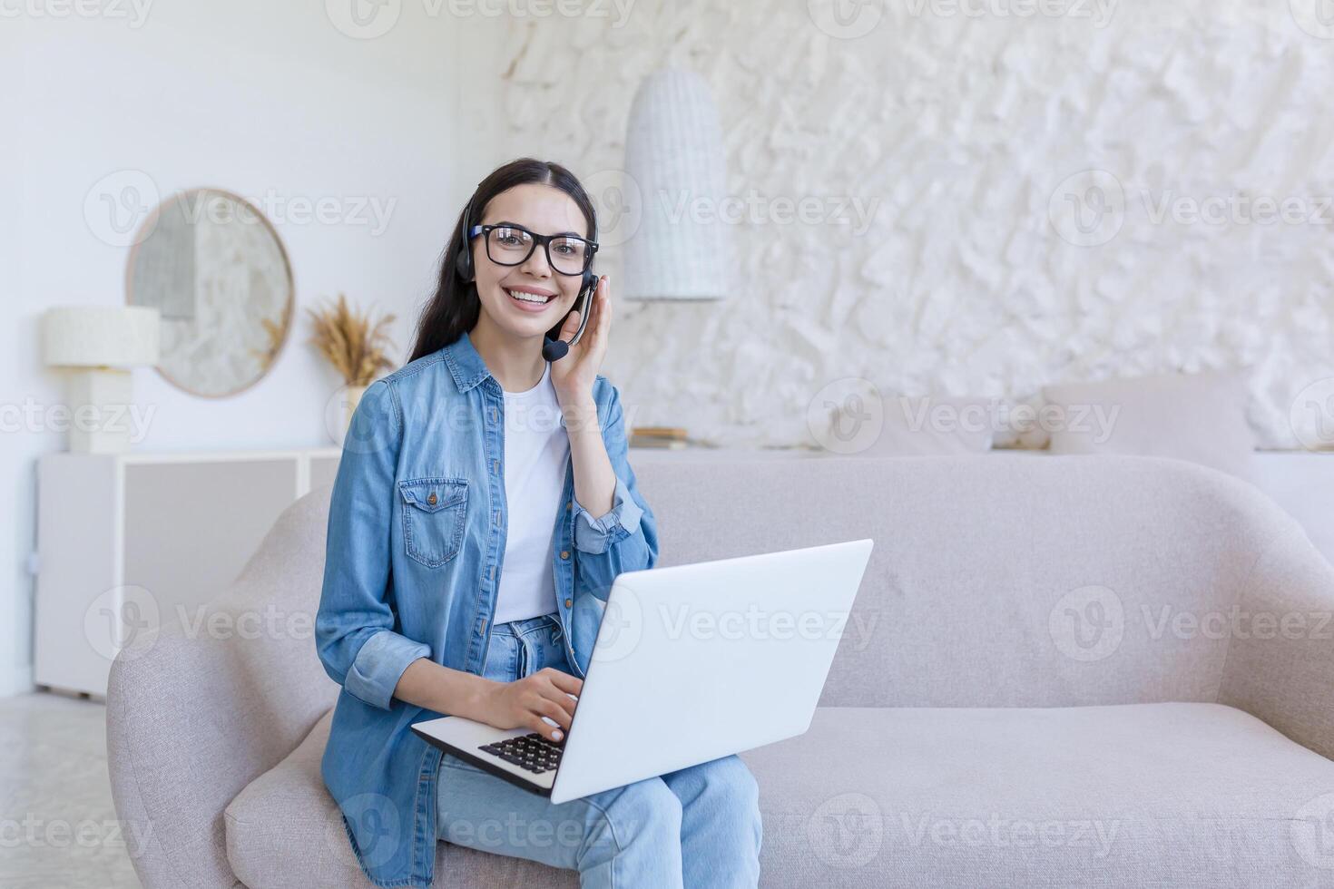 Portrait of a beautiful young female student sitting on the sofa at home with a laptop and a headset. He studies online, remotely. He looks at the camera, smiles. photo