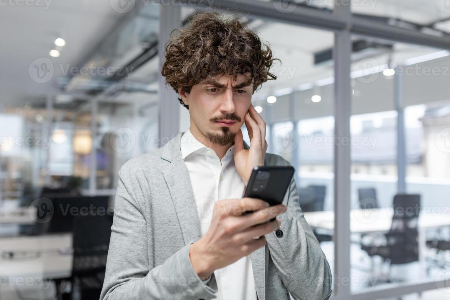 Businessman in office near window upset reading message online notification from phone, male boss working inside office in business suit with beard, upset investor. photo