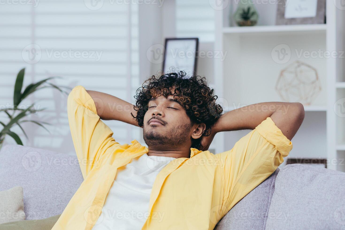 Young man relaxing alone at home on sofa, close-up Hispanic man with hands behind head dozing and dreaming with closed eyes in living room of house. photo
