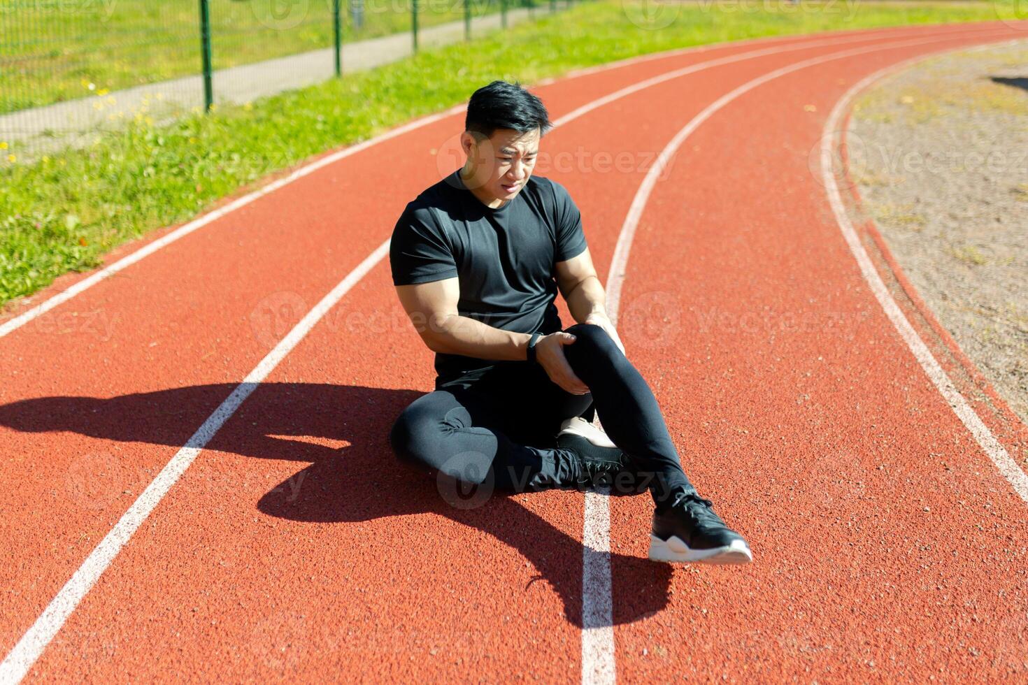 Sports injury. A young Asian sportsman, athlete, runner sits on a stadium treadmill and holds his leg, knee. feels severe pain. photo